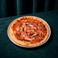 Cooked Doughvinci Meatlovers pizza topped with salami, ham, tomato sauce and melted cheese on a blistered woodfired sourdough crust, served on a white plate on a dark green velvet table with matching curtains behind