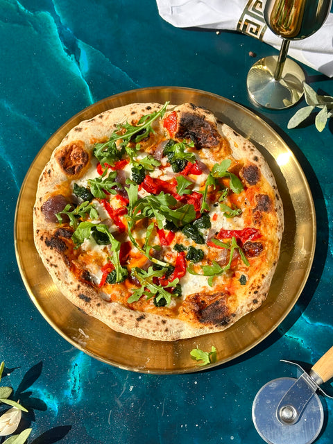 A round pizza topped with arugula, red peppers, and melted cheese, served on a round gold platter over a teal surface with a wine glass and pizza cutter nearby.